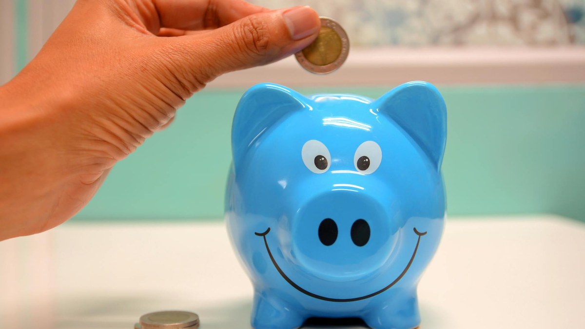 close-up of a hand placing coins into a clear piggy bank on a wooden desk