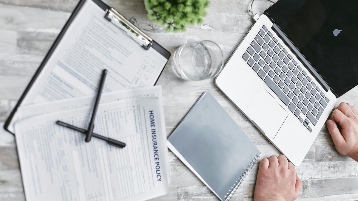 Person thoughtfully reviewing insurance documents and a laptop with financial charts on screen